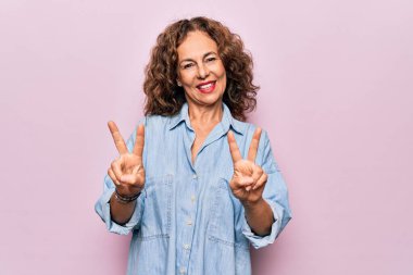 Orta yaşlı güzel bir kadın pembe arka planda dikilmiş sıradan bir kot gömlek giyiyor ve iki elini de zafer işareti yaparken göstererek gülümsüyor. İki numara..