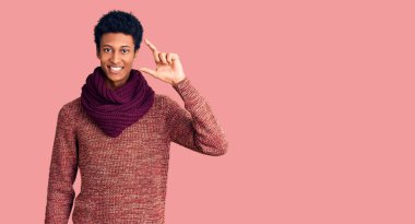 Genç Afro-Amerikan adam günlük kışlık süveter ve eşarp giyiyor gülümsüyor ve kendine güvenen el hareketleriyle küçük beden işareti yapıyor parmaklarıyla ve kamerayla bakıyor. ölçüm kavramı. 