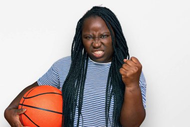 Saçları örgülü, elinde basketbol topu olan genç siyahi bir kadın öfkeyle bağırıyor, öfkeyle bağırıyor ve el kaldırıyor. 