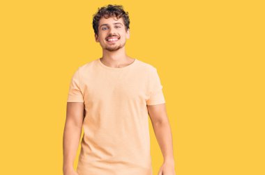 Kıvırcık saçlı, sıradan giyinen, pozitif ve mutlu görünen, kendine güvenen gülümseyen, dişlerini gösteren genç ve yakışıklı bir adam. 