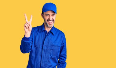 Orta yaşlı, tamirci üniforması giyen, gülümseyen ve kameraya göz kırpan yakışıklı bir adam parmaklarıyla zafer işareti yapıyor. İki numara.. 