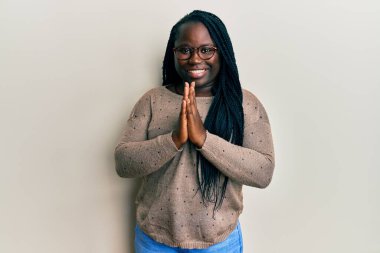 Saçları örgülü, sıradan giyinmiş, gözlüklü, elleri kollarında dua eden, affedilmek isteyen, kendinden emin gülümseyen genç siyahi bir kadın.. 