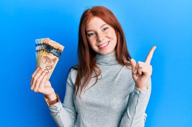 Kanada doları tutan genç kızıl saçlı kız mutlu bir şekilde gülümsüyor. 