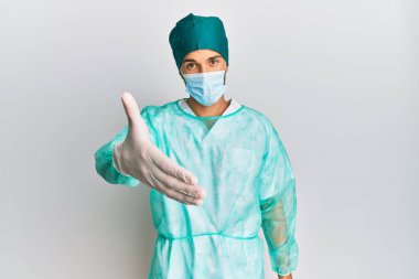 Cerrah üniforması ve tıbbi maske takan genç yakışıklı adam dostça bir selamlama ve hoş geldin hediyesi olarak el sıkışmayı sunuyor. başarılı iş. 