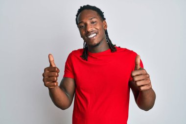 Saçları örgülü Afro-Amerikan bir adam günlük kıyafet giyer başarı işareti yapar, elleriyle olumlu jest yapar, gülümser ve mutlu olur. Neşeli ifade ve kazanan jest. 