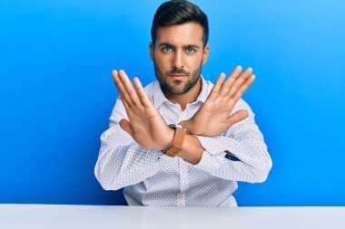 Yakışıklı İspanyol bir adam iş kıyafetleri içinde masada oturuyor. Reddedilmiş yüz ifadesi kollarını kavuşturuyor. Negatif işaretler yapıyor. 