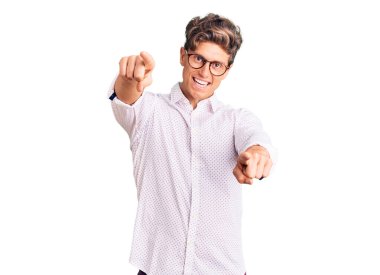 İş kıyafetleri ve gözlük takan genç yakışıklı adam seni ve kamerayı işaret ediyor, pozitif ve neşeli gülümsüyor. 