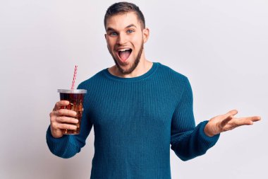 Young handsome man dirnking cola refreshment beverage celebrating achievement with happy smile and winner expression with raised hand 