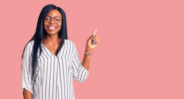 Genç Afrikalı Amerikalı kadın, günlük kıyafetler ve gözlük takıyor. Yüzünde kocaman bir gülümseme var. Elinle kameraya bakıyor.. 