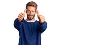 Sakallı, sıradan bir süveter giyen, elinde olumlu bir jest yapan, gülümseyen ve başarı için sevinen yakışıklı sarışın bir adam. kazanan jest. 