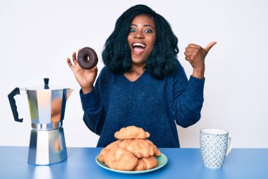 Güzel bir Afrikalı kadın kahvaltısını yapıyor. Çikolatalı donut tutuyor. Parmağını kaldırıp gülümsüyor. 