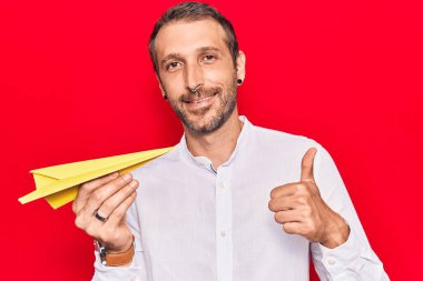 Elinde kağıt uçak tutan yakışıklı adam mutlu ve pozitif gülümsüyor, başparmağıyla mükemmel ve onay işareti yapıyor. 