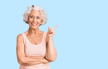 Yaşlı, gri saçlı, günlük kıyafetler giyen, gülümseyen mutlu, eli ve parmağı yanda olan bir kadın. 