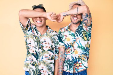 İki genç eşcinsel adam yaz şapkası takıyor ve Hawaii gömleği giyip gözleri kollarıyla kaplıyor. Neşeli ve komik gülümsüyorlar. kör kavram. 