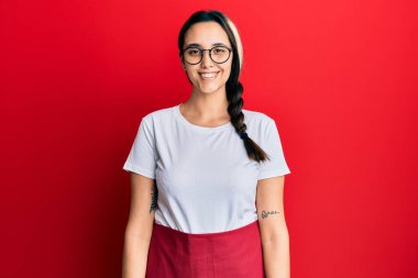 Genç İspanyol kadın, yüzünde mutlu ve havalı bir gülümsemeyle profesyonel garson önlüğü giyiyor. şanslı kişi. 