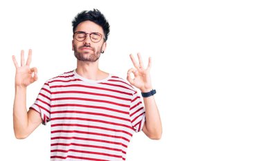 Gündelik kıyafetler giyen, gözleri kapalı gülümseyen genç İspanyol adam parmaklarıyla meditasyon hareketi yapıyor. yoga konsepti. 