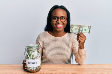 Genç Afrikalı bir kadın elinde dolar ve kavanozla pozitif ve mutlu görünüyor ve kendine güvenen bir gülümsemeyle dişlerini gösteriyor. 