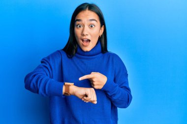 Kış süveteri giymiş genç Asyalı bir kadın acele edip saati, sabırsızlığı ve gecikmeye kızmayı işaret ediyor. 