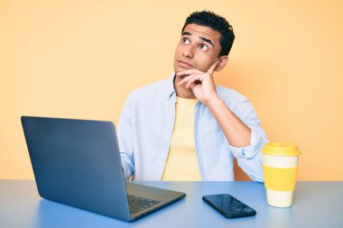 Young handsome hispanic man working at the office with laptop serious face thinking about question with hand on chin, thoughtful about confusing idea 