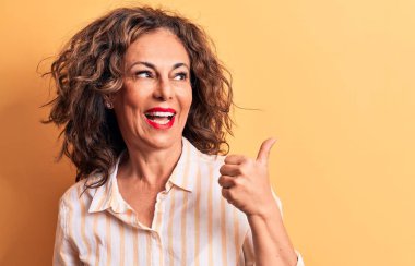 Orta yaşlı, esmer, çizgili tişörtlü, sarı arka planda duran güzel bir kadın baş parmağını yukarı kaldırıyor ve ağzı açık gülümsüyor.