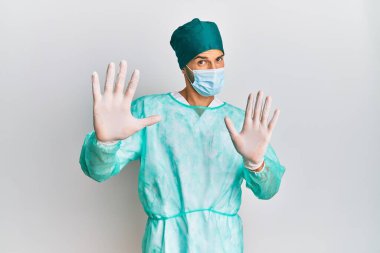 Cerrah üniforması ve tıbbi maske takan genç yakışıklı adam korku ifadesinden korkuyor ve dehşete düşüyor. El hareketlerini durdur, şok içinde bağır. panik konsepti. 