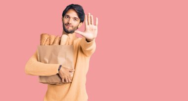 Yakışıklı İspanyol bir adam elinde bir kese kağıdıyla elinde ekmekle dur işareti yapıyor ciddi ve kendinden emin bir ifadeyle, savunma jesti yapıyor. 