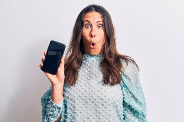 Genç, güzel, esmer bir kadın elinde akıllı telefonuyla beyaz arka planda korku ve şaşkınlık içinde şaşkınlık dolu bir ifade veriyor.