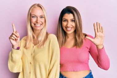 Pembe arka planda yan yana duran iki arkadaş altı numaralı parmaklarını gösterip işaret ederken kendinden emin ve mutlu gülümsüyor.. 