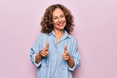 Orta yaşlı güzel bir kadın pembe arka planda dikilmiş sıradan bir kot gömlek giyiyor ve mutlu ve komik yüzlü parmakları kameraya doğrultuyor. İyi enerji ve titreşimler.