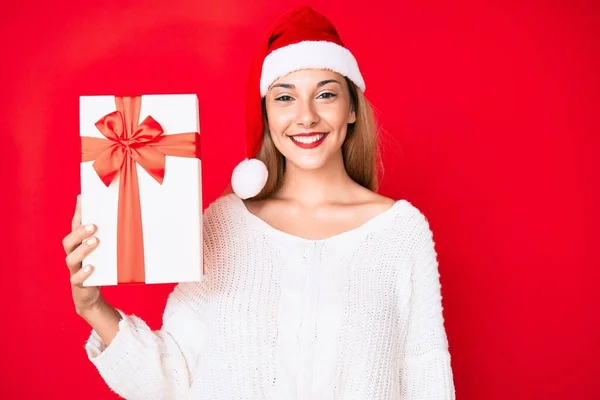 Young Brunette Woman Wearing Christmas Hat Holding Gift Looking Positive — Stock fotografie