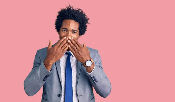 Handsome african american man with afro hair wearing business jacket laughing and embarrassed giggle covering mouth with hands, gossip and scandal concept