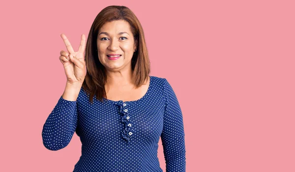 Orta yaşlı Latin kadın sıradan kıyafetler giyip kameraya gülümseyen ve zafer işareti yapan. İki numara.. 
