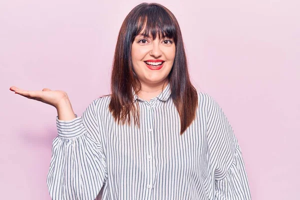 Genç ve cüsseli bir kadın günlük kıyafetler giyiyor neşeli bir şekilde gülümsüyor ve avuç içi kameraya bakıyor.. 