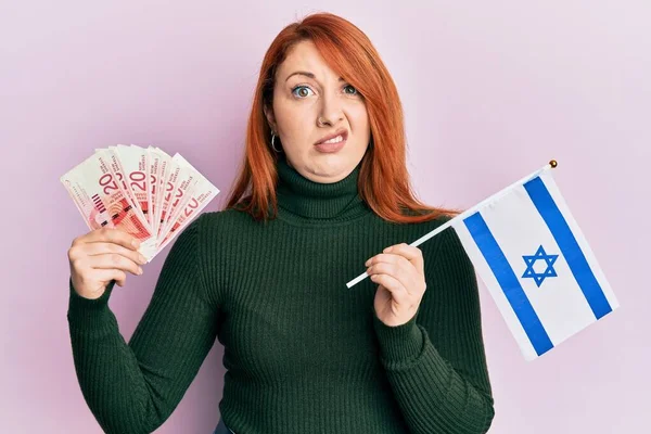 漂亮的红头发女人手里拿着20谢克尔钞票和以色列国旗 毫无头绪 表情混乱 怀疑概念 — 图库照片