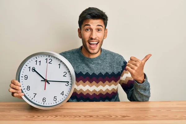 Joven Hombre Guapo Sosteniendo Gran Reloj Apuntando Con Pulgar Hacia —  Fotos de Stock