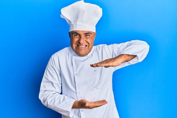 プロの料理の制服を身に着けている成熟した中東の男性と手でジェスチャーで大小のサインを示す帽子は シンボルを測定します カメラを見て笑ってる 概念を測定し — ストック写真