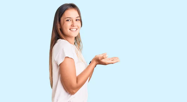 コピースペースを示す手を開いて手のひらで脇を指すカジュアルな白いTシャツを身に着けている美しい白人女性 広告を提示笑顔興奮幸せ — ストック写真