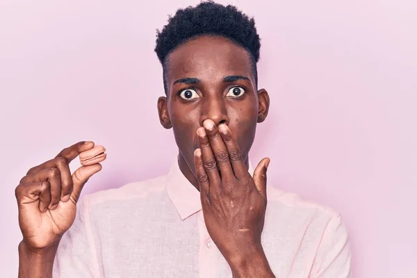 Joven Afroamericano Sosteniendo Macaron Cubriendo Boca Con Mano Sorprendido Temeroso — Foto de Stock