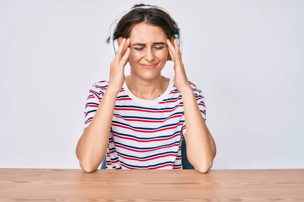 年轻的惊慌失措的女人用耳机坐在桌子上 由于疼痛和偏头痛 绝望和压力重重 手放在头上 — 图库照片