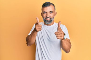 Orta yaşlı beyaz tişört giyen yakışıklı bir adam mutlu ve komik yüzüyle kamerayı işaret ediyor. iyi enerji ve titreşimler. 
