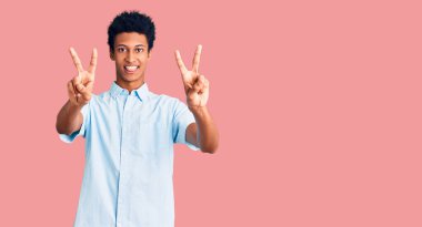 Gündelik giysiler giymiş genç Afro-Amerikan bir adam iki elini de zafer işareti yaparken göstererek gülümsüyor. İki numara.. 