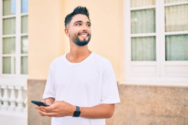 Genç Arap adam şehirde akıllı telefon kullanarak mutlu gülümsüyor..