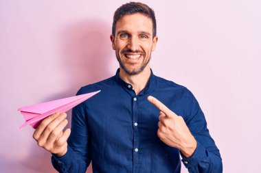 Pembe kağıt uçak tutan genç yakışıklı adam izole bir arka planda duruyor ve mutlu bir şekilde elini ve parmağını göstererek gülümsüyor.