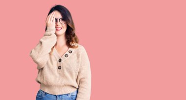 Güzel, genç, esmer bir kadın günlük kıyafetler ve gözlük takıyor bir gözünü eliyle kapatıyor, yüzünde kendine güvenen bir gülümseme ve sürpriz bir duygu var.. 