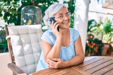 Gri saçlı, gülümseyen, mutlu, rahatlatıcı orta yaşlı bir kadın terasta oturmuş telefonla konuşuyor.