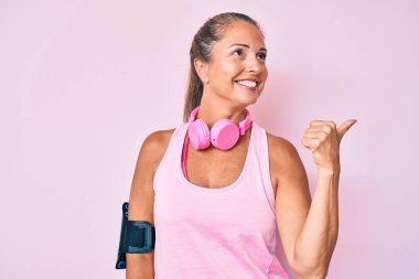 Orta yaşlı İspanyol bir kadın spor kıyafetleri giyiyor ve kulaklık takıyor. Gülümsüyor, gülümsüyor ve baş parmağıyla yan tarafa bakıyor.. 