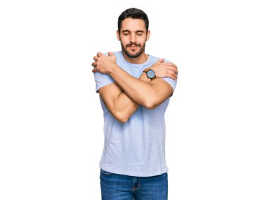 Gündelik kıyafetler giyen genç İspanyol adam kendine sarılıyor mutlu ve pozitif, kendinden emin gülümsüyor. Sevgi ve özgüven. 