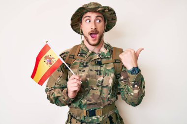Kamuflaj üniforması giyen genç beyaz adam elinde İspanyol bayrağı başparmağıyla yan tarafa bakıyor ve ağzı açık gülümsüyor. 