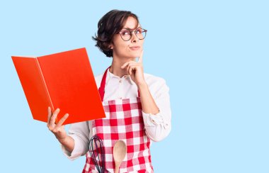 Kısa saçlı, profesyonel fırıncı önlüğü giyen güzel bir kadın yemek tarifi kitabı okuyor ciddi bir yüz ifadesi var eli çenesinde soru soran, kafa karıştırıcı bir fikir hakkında düşünceli. 