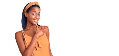 Genç Afro-Amerikan kadın, yaz kıyafetleri içinde neşeli ve yüzünde mutlu ve doğal bir ifadeyle, elleri ve parmaklarıyla yüzünü gösteren bir gülümseme ile. 